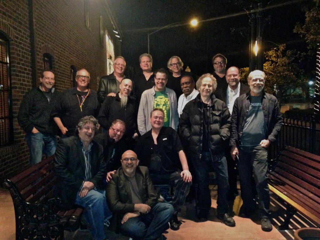 A group of sixteen men pose together outside at night, some standing and some sitting on benches, in front of a brick building with streetlights and trees visible in the background.