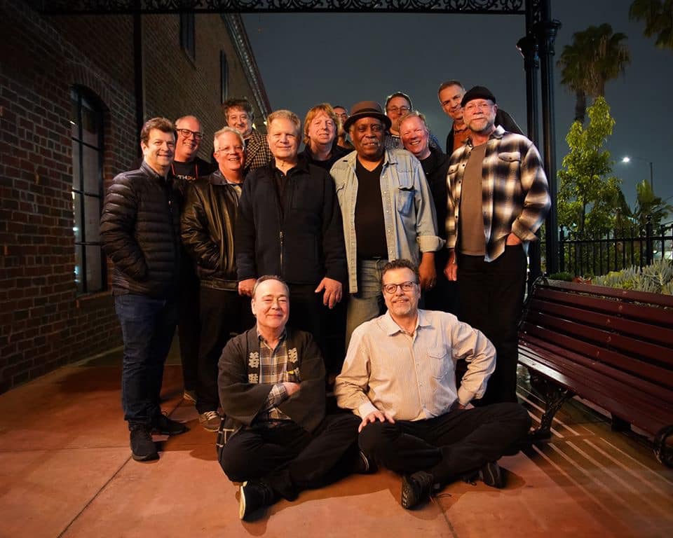 A group of fifteen men pose together outdoors at night, some standing and some sitting on the ground, in front of a brick building and iron fence with trees and streetlights in the background.