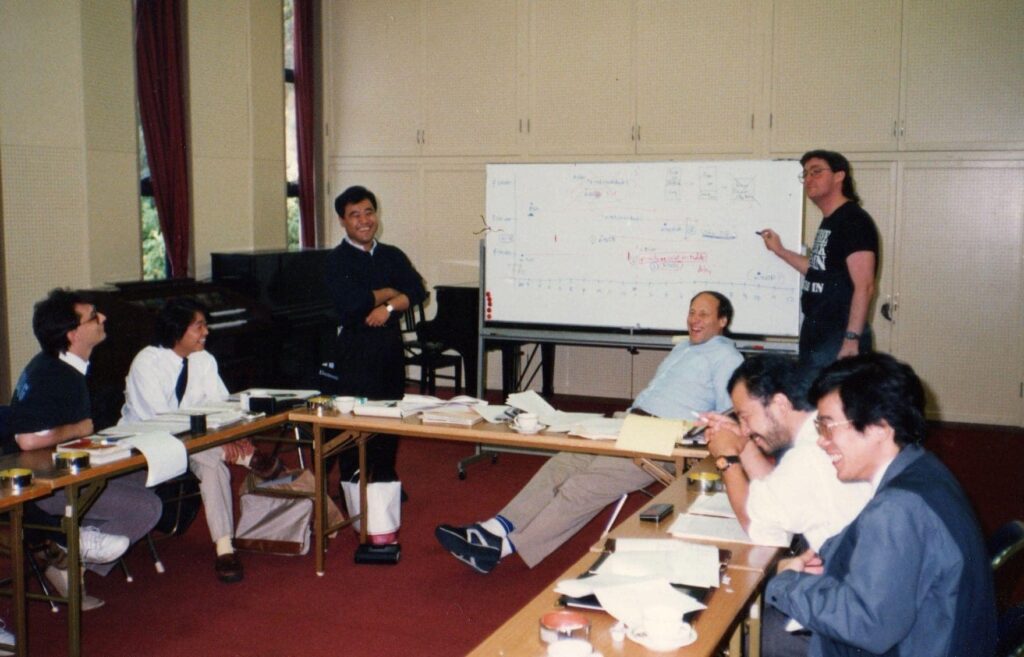 A group of seven people sit and stand around tables in a classroom. One person stands at a whiteboard with diagrams and notes, while others are seated, talking or laughing, with papers and folders on the tables.
