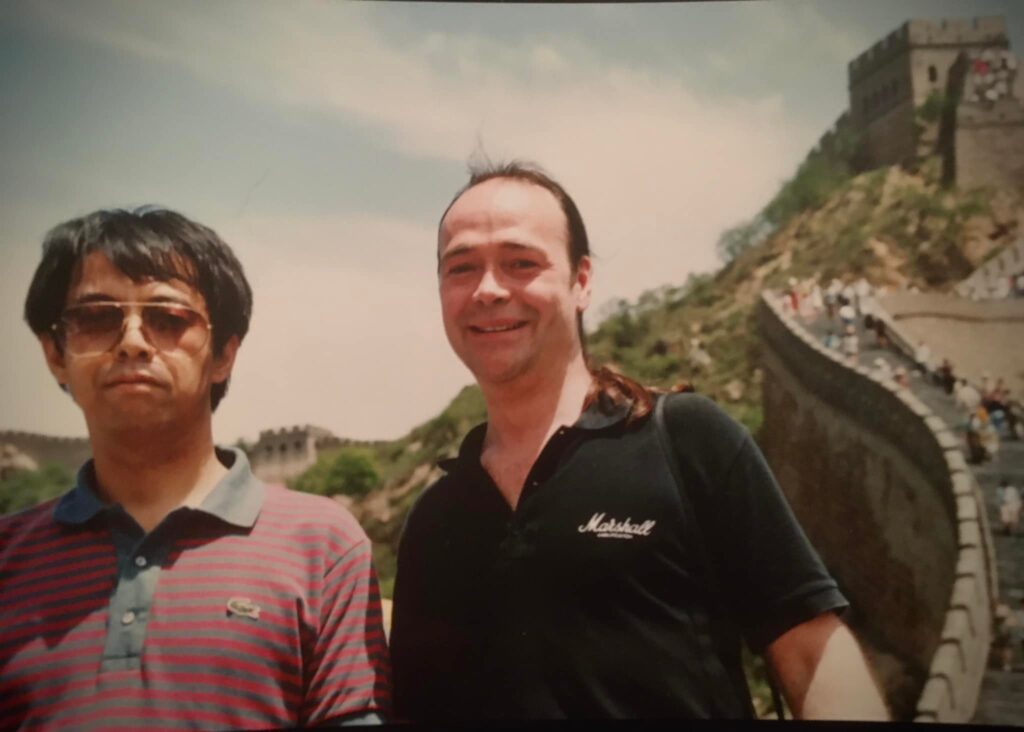 Rio Takahashi and Jack Hotop from Korg stand on the Great Wall of China. One wears sunglasses and a striped polo shirt; the other wears a black shirt and smiles. The wall curves along a green hillside under a sunny sky, with other visitors in the background.