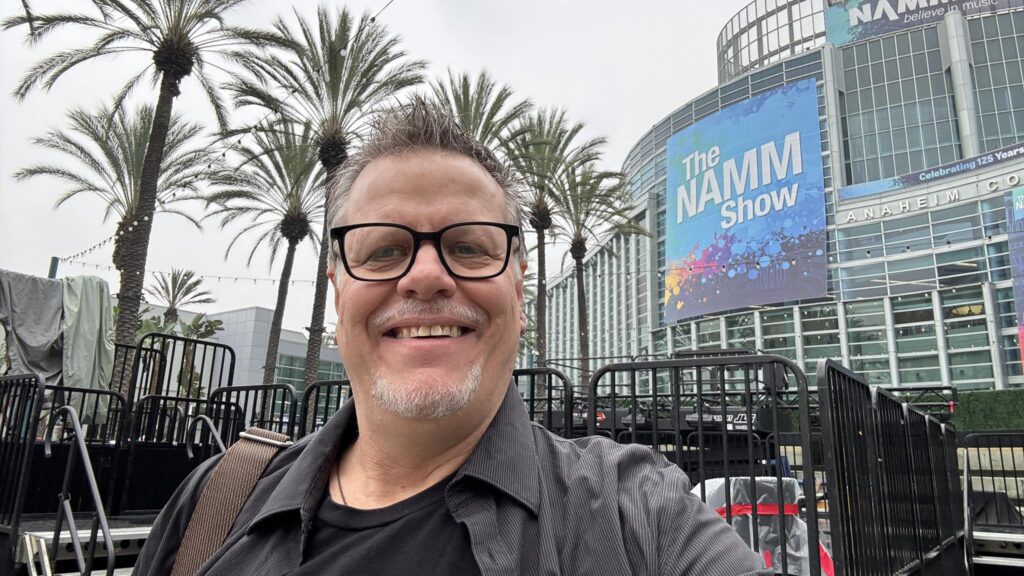 A smiling man with glasses takes a selfie outside the Anaheim Convention Center, with palm trees and a large blue The NAMM Show sign visible in the background.