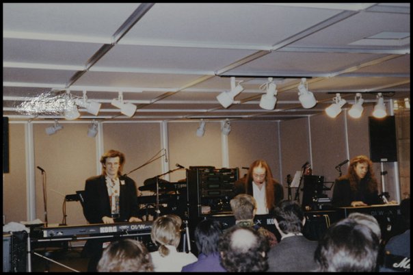 Three musicians play keyboards on a stage in front of an audience. The room has modern lighting, sound equipment, and a neutral-colored ceiling and walls. The audience is seated, watching the performance.