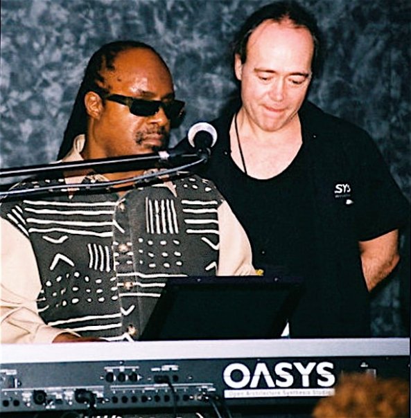 Jack and Stevie Wonder stand behind an OASYS keyboard; the man on the left wears sunglasses and a patterned shirt, playing the keyboard, while the man on the right, in a black shirt, looks on and smiles.