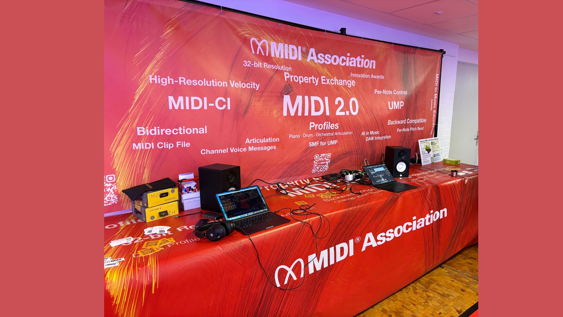 A display booth for the MIDI Association features a red banner with white text about MIDI 2.0, technology terms, and logos. The table holds laptops, audio equipment, and cables in a brightly lit room.