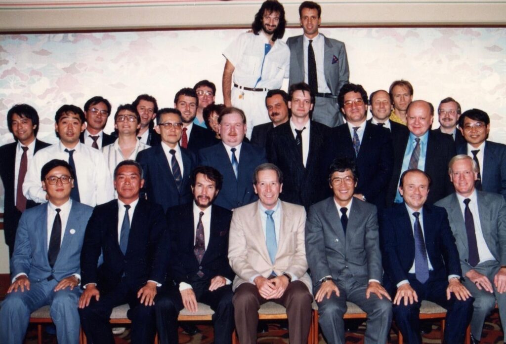 A group of about 27 men in formal and business attire pose together indoors, arranged in four rows, with some seated in front and others standing behind them. The background features a pale, patterned wall.
