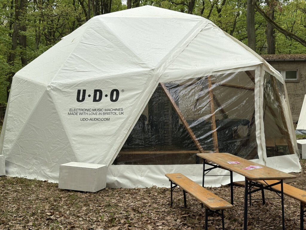 A white geodesic dome tent with clear plastic windows sits in a forest. The dome displays U-D-O Electronic Music Machines Made With Love in Bristol, UK. Wooden benches and tables are set up outside on the leaf-covered ground.