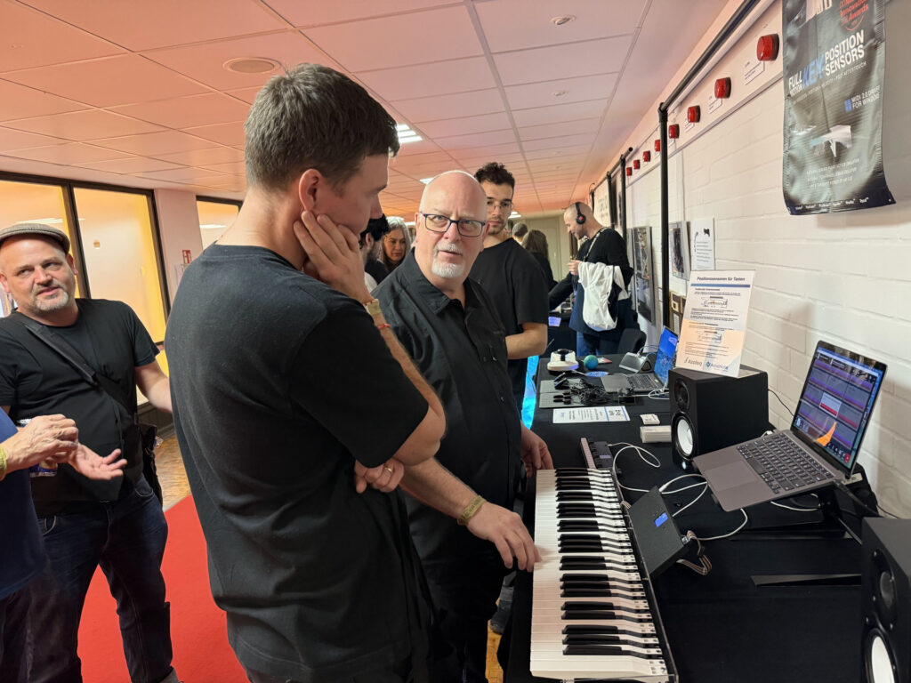 A group of people stands around a digital keyboard and music production equipment in a hallway. Two men in the center discuss the setup, while others observe. Posters are visible on the white brick wall behind them.