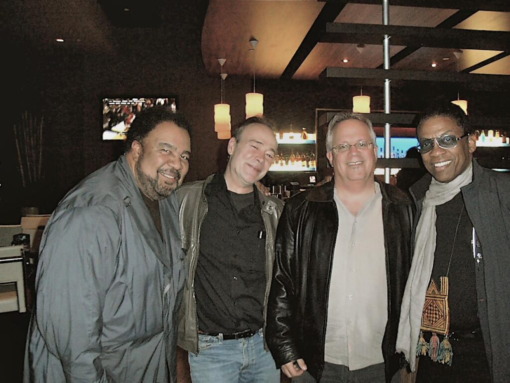 George Duke, Jack, Jerry Kovarsky and Herbie Hancock stand together smiling in a dimly lit bar or lounge, dressed in casual jackets. Shelves with bottles and hanging lights are visible in the background.