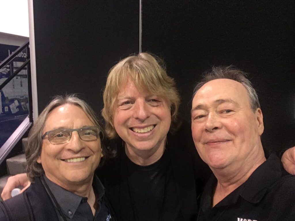 Three smiling men stand close together for a selfie, with two wearing glasses and collared shirts and the man in the center with blond hair. They are indoors in front of a black divider.
