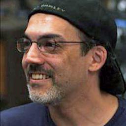 A man with glasses, a black baseball cap worn backwards, and a short beard is smiling and looking to the side. He is wearing a dark shirt.