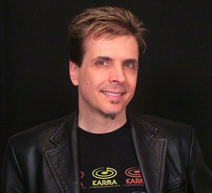 A man with short, light brown hair and a trimmed goatee is smiling. He is wearing a black leather jacket over a black T-shirt with colorful designs and the word KARMA repeated. The background is plain black.