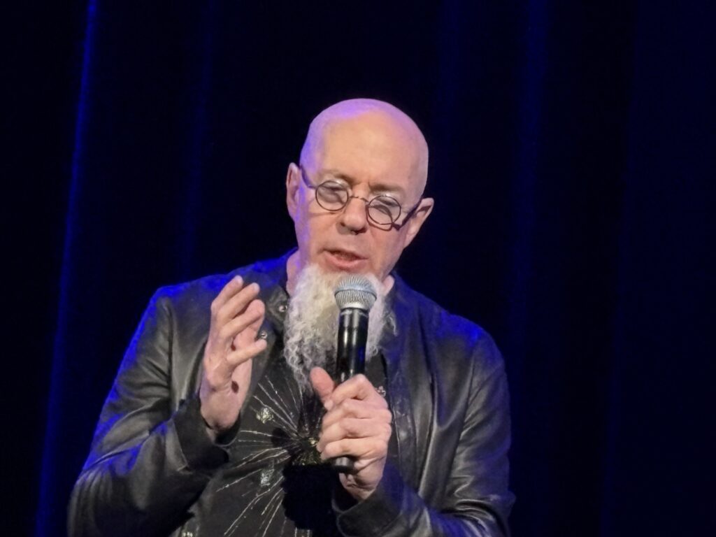Jordan Rudes with round glasses and a long white goatee speaks into a microphone on stage, wearing a black leather jacket, with a dark blue curtain in the background.