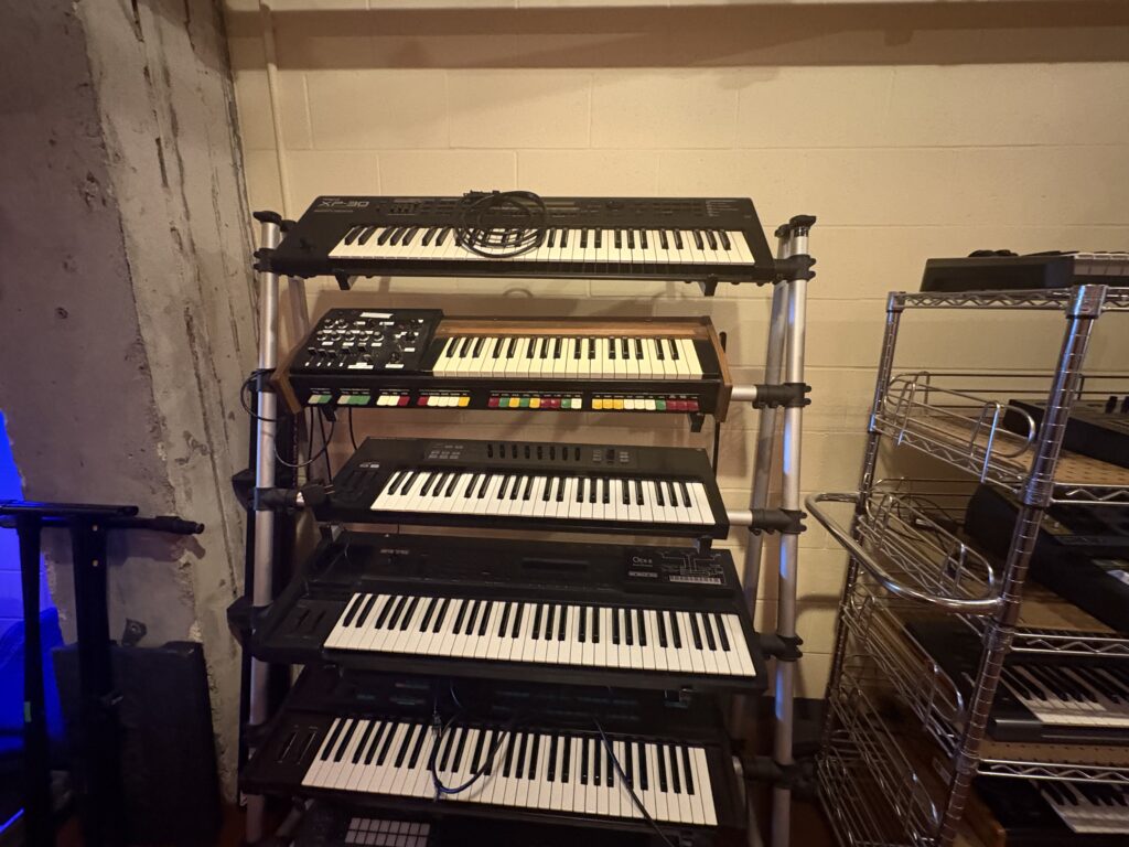 A set of five electronic keyboards and synthesizers are stacked on a multi-tiered metal rack against a beige wall, with some cables resting on the top keyboard. An empty wire shelving unit is on the right.