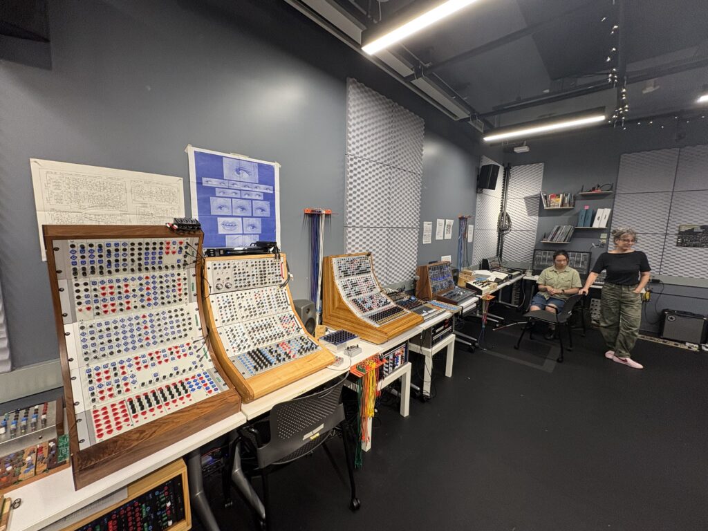 A music studio with modular synthesizers on desks, soundproofing panels on gray walls, and two people standing and sitting near shelves with equipment and books. The room has bright ceiling lights.