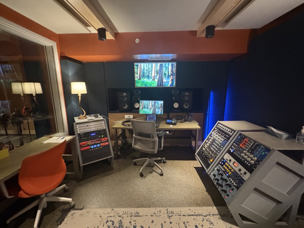 A modern audio production studio with two desks, computer monitors displaying a forest scene, audio equipment racks, studio speakers, and chairs. The room has orange and black walls, and acoustic treatment on the ceiling.
