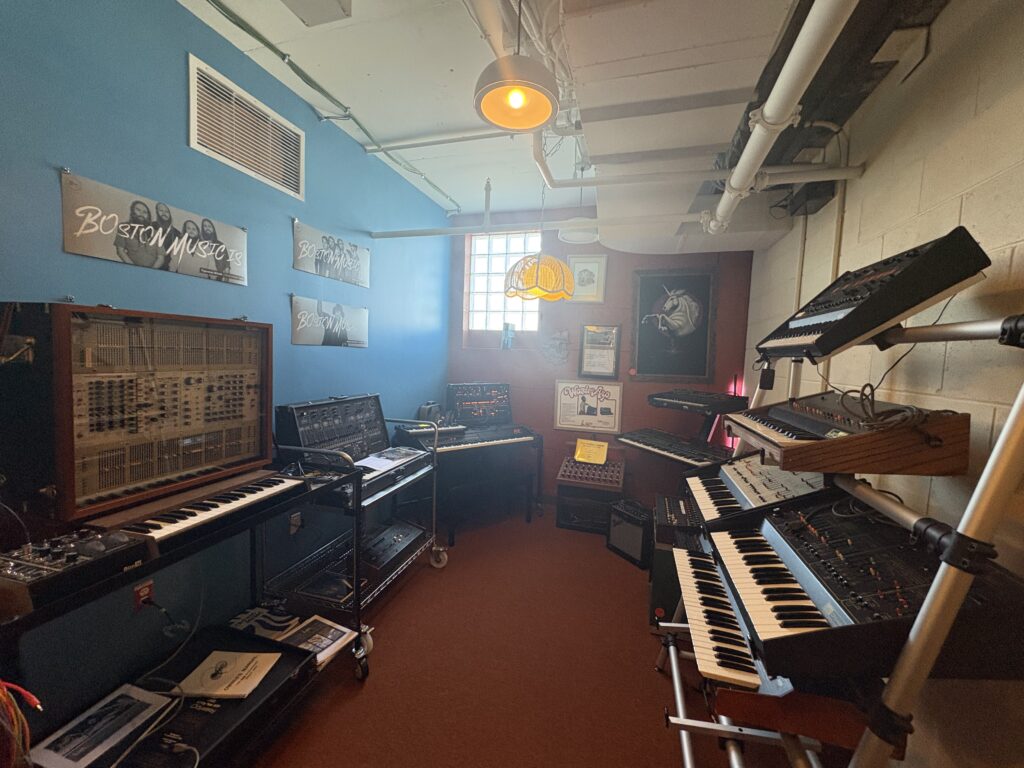 A small room with blue walls features several vintage synthesizers on shelves, a hanging lamp, posters on the walls, and a window letting in daylight, creating a cozy music studio atmosphere.