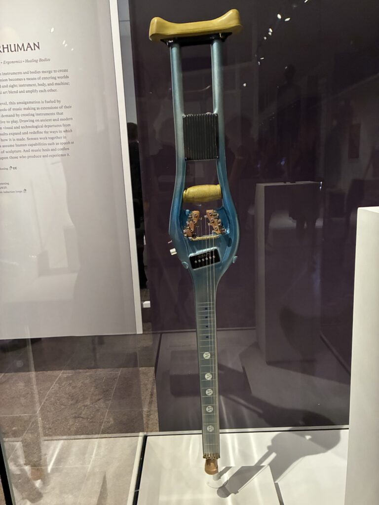 A guitar shaped like a crutch with strings, tuning pegs, and internal electronic components is displayed upright in a museum case. The crutchs handle and padding are visible at the top.