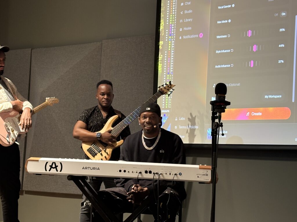 Three musicians in a studio, one playing bass guitar, another sitting at an Arturia keyboard, and a third with a guitar. Music software is projected on the wall behind them.