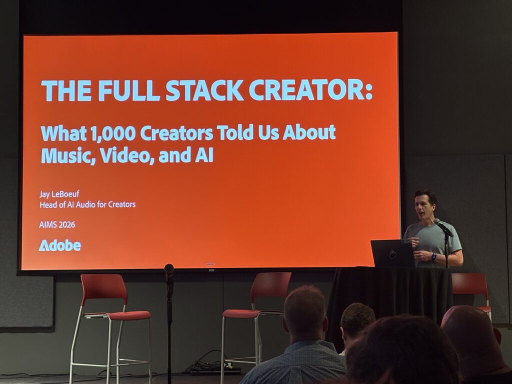 Jay LeBeouf from Adobe stands at a podium beside a large screen displaying a presentation titled THE FULL STACK CREATOR: What 1,000 Creators Told Us About Music, Video, and AI by Jay LeBoeuf from Adobe, with an audience seated in front.