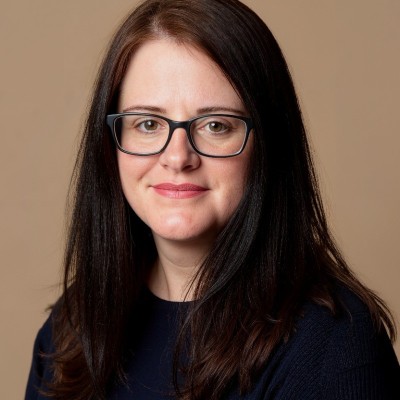 A woman with straight, dark brown hair and glasses is smiling softly. She is wearing a dark top and is posed in front of a plain beige background.