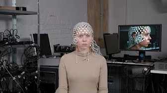 A person wearing an EEG cap sits in a lab with electronic equipment and cables. A computer monitor behind them displays their side profile, also wearing the cap.