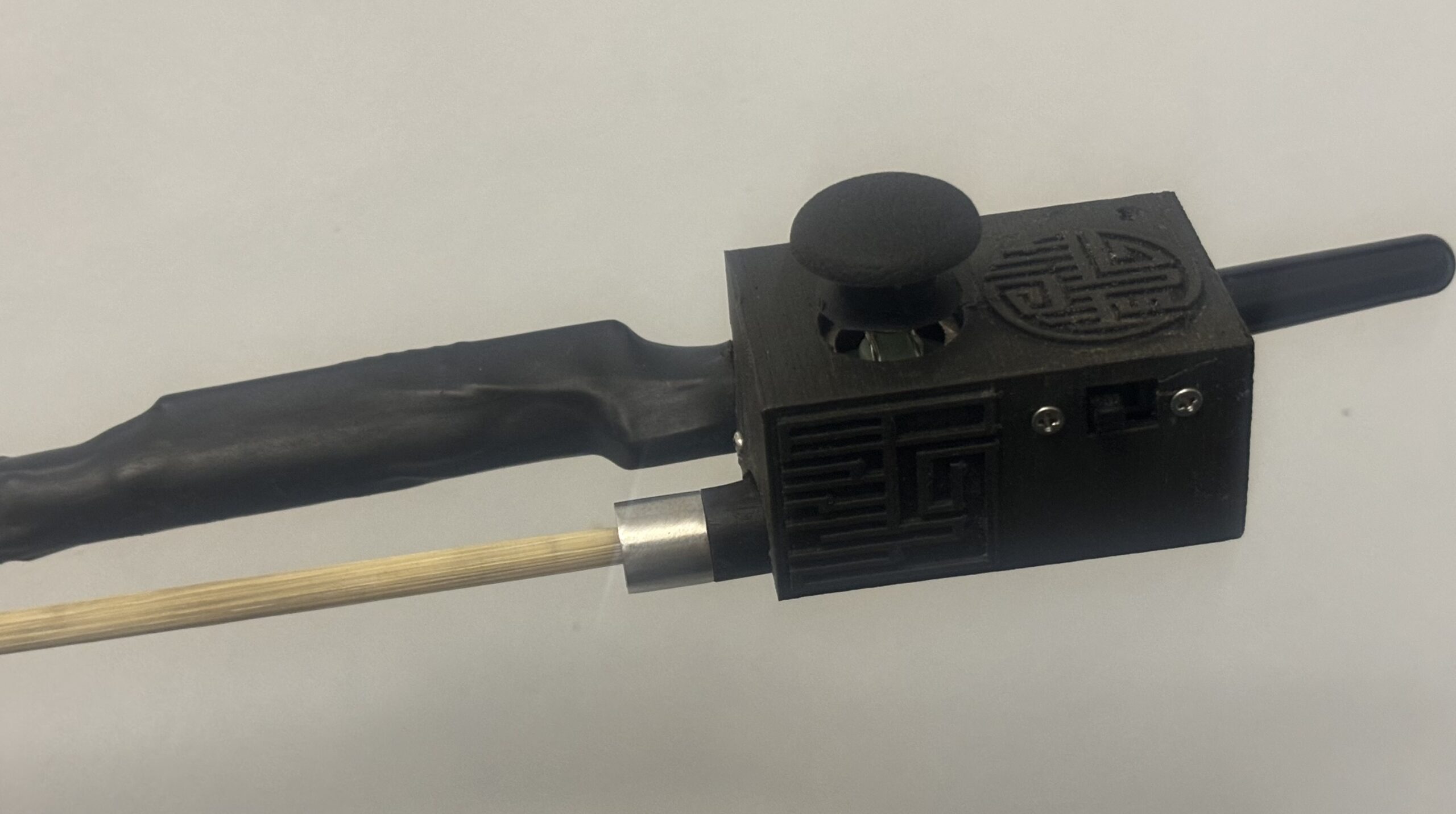 A close-up of a device attached to a wooden stick, featuring a textured grip, a round black knob, and decorative geometric patterns on a rectangular black box with a switch.