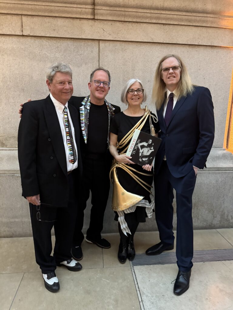 Four adults in formal attire stand together, smiling for the camera. One woman holds a book; two men wear suits and one wears a jacket with a piano-key design. They are standing against a stone wall indoors.