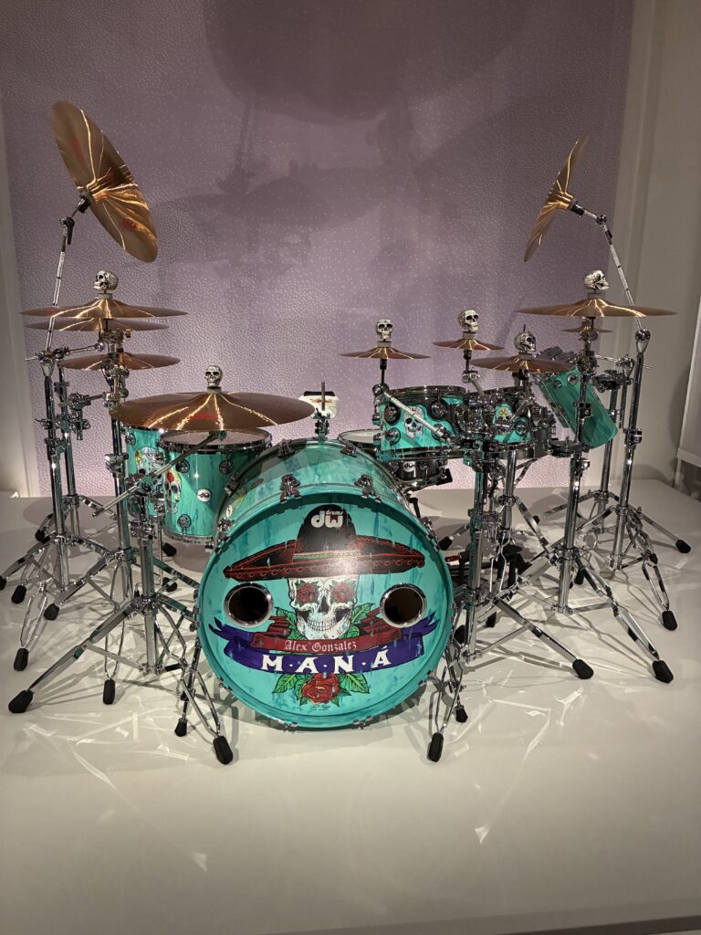 A turquoise drum set with skull-shaped decorations on the hardware, featuring artwork and the word “Maná” on the bass drum, displayed on a white platform against a light textured background.