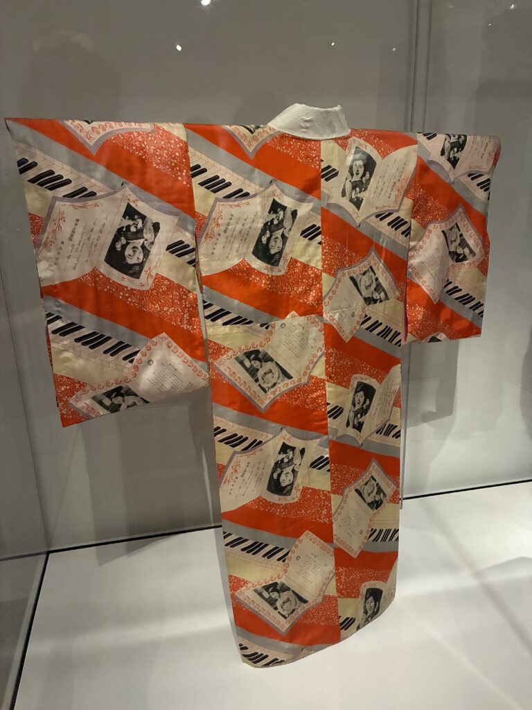 A colorful kimono displayed in a glass case, featuring a repeating pattern of photographs, handwritten notes, and musical staff designs on a red, white, and beige background.