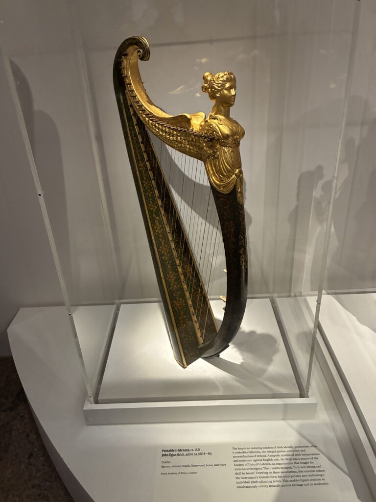 A decorative harp with a gold figure of a woman at the top stands in a glass display case on a white platform; information about the instrument is shown on a plaque below.