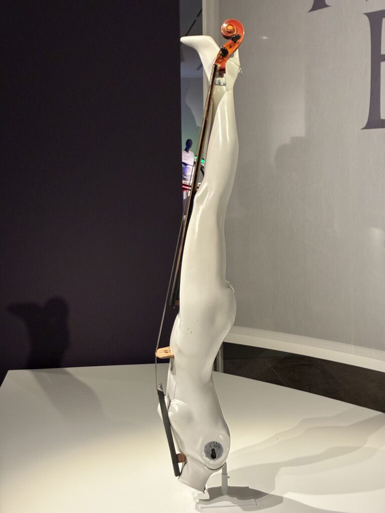 A sculptural artwork shaped like an upside-down human figure, painted white, with a violin integrated along its back, displayed on a white pedestal against a dark background.
