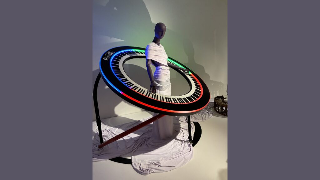 A mannequin wrapped in white fabric stands inside a large, circular keyboard with colorful lights, resembling a futuristic piano. The keyboard is tilted and supported by black stands; draped fabric pools at the base.