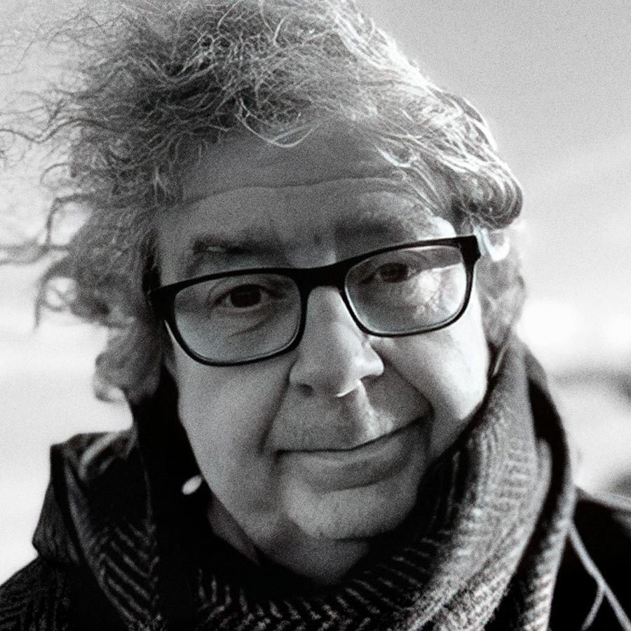 A black-and-white photo of an older person with curly, windblown hair, wearing dark-framed glasses and a scarf, looking at the camera with a gentle expression.