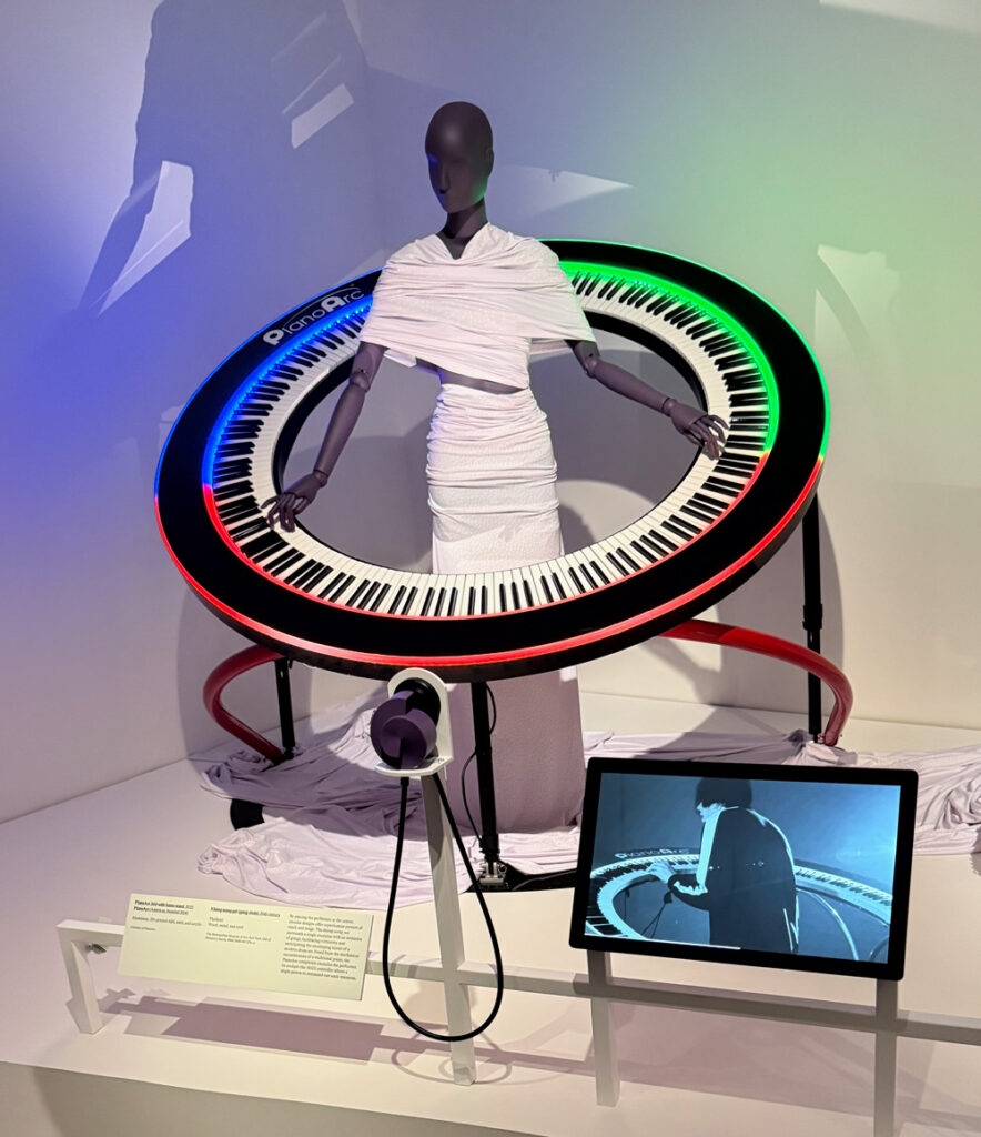 A mannequin in a white wrap dress stands inside a large circular keyboard with colored LED lights; a screen nearby displays a video of a performer using the same instrument.