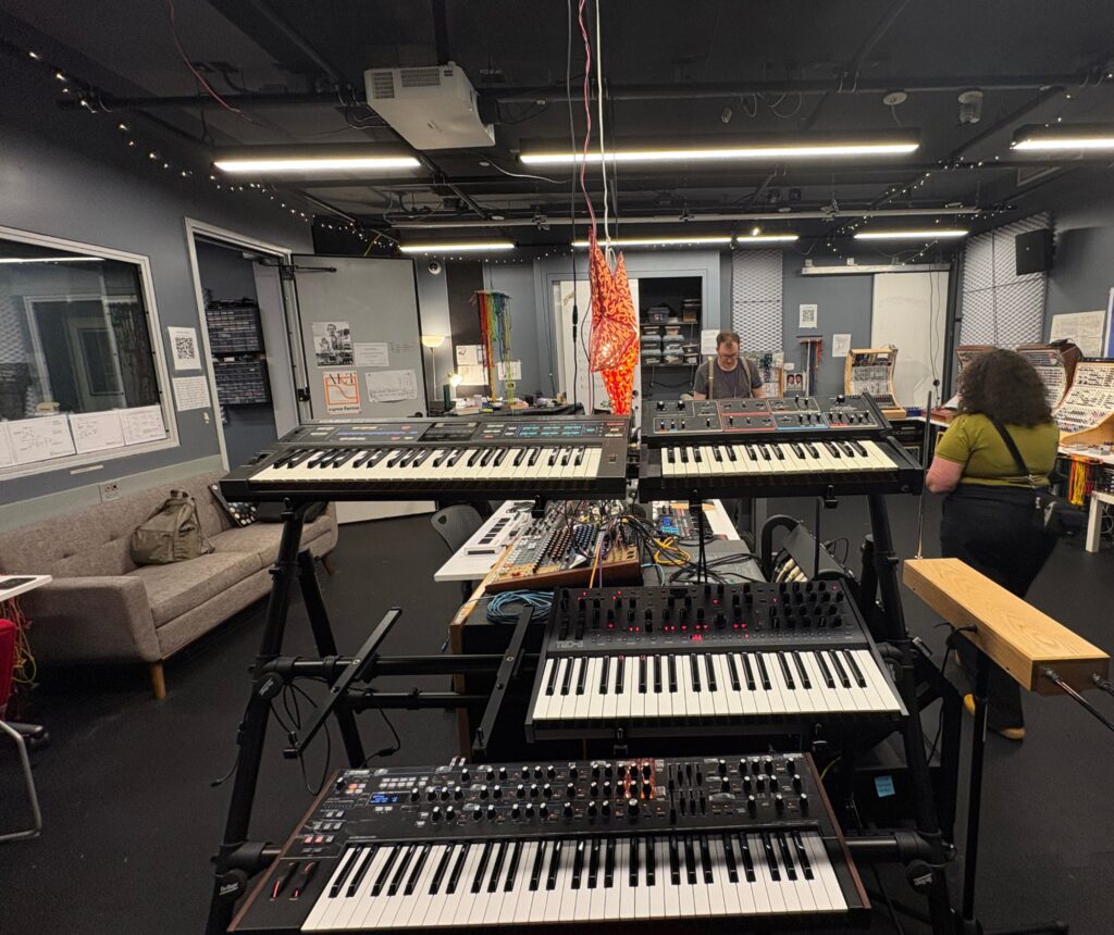 A music studio with multiple synthesizers on stands, cables, a modular synth setup, two people talking, a sofa, shelves with equipment, and posters on the walls. Warm lighting and a relaxed atmosphere.