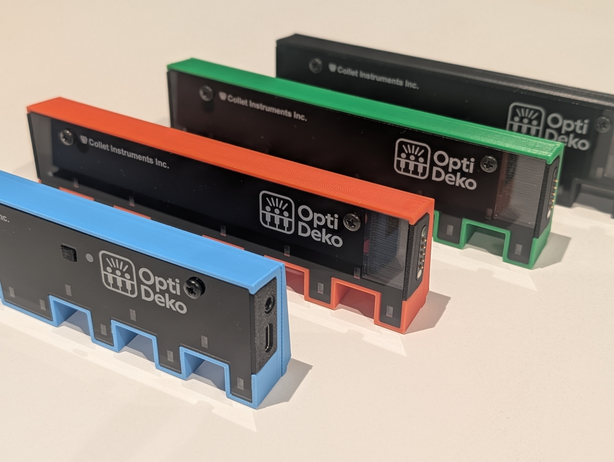 Four rectangular electronic devices labeled Opti Deko in black, each with different colored bases (blue, orange, green, and white), are arranged in a diagonal line on a light surface.