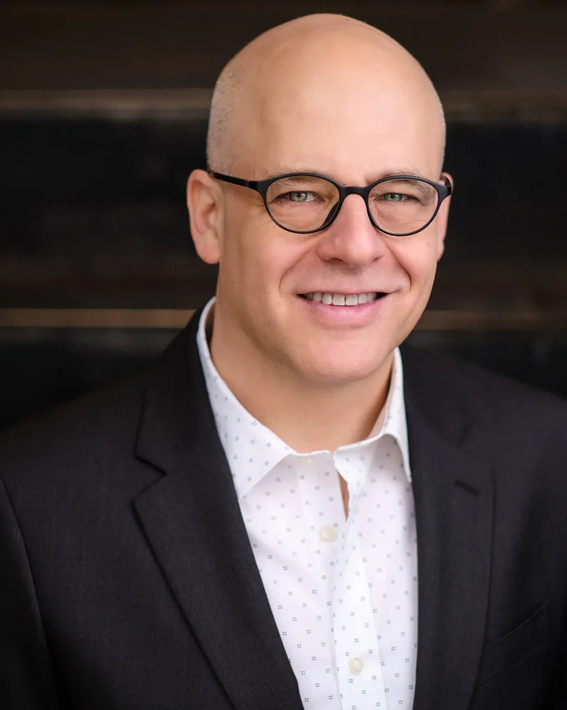 A smiling man wearing glasses, a dark suit jacket, and a white patterned shirt, posing in front of a dark, blurred background.