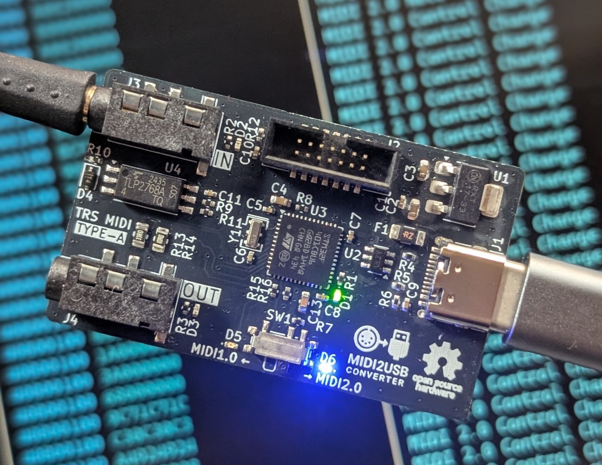 A close-up of a MIDI2USB converter circuit board connected by USB-C, with several electronic components visible, lying on a surface with a blurred blue and black digital pattern in the background.