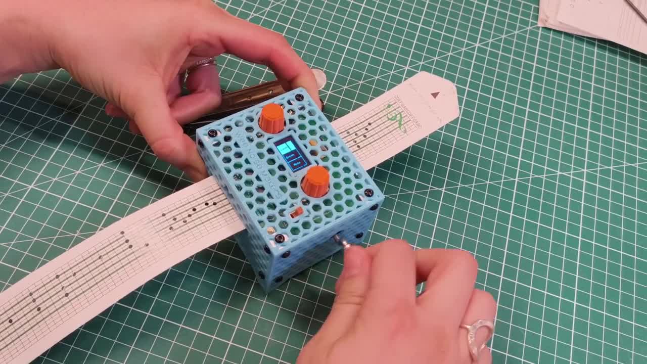 A person uses a blue, perforated electronic device with orange dials and a small display to scan or read a strip of paper with printed dots and symbols on a green cutting mat.