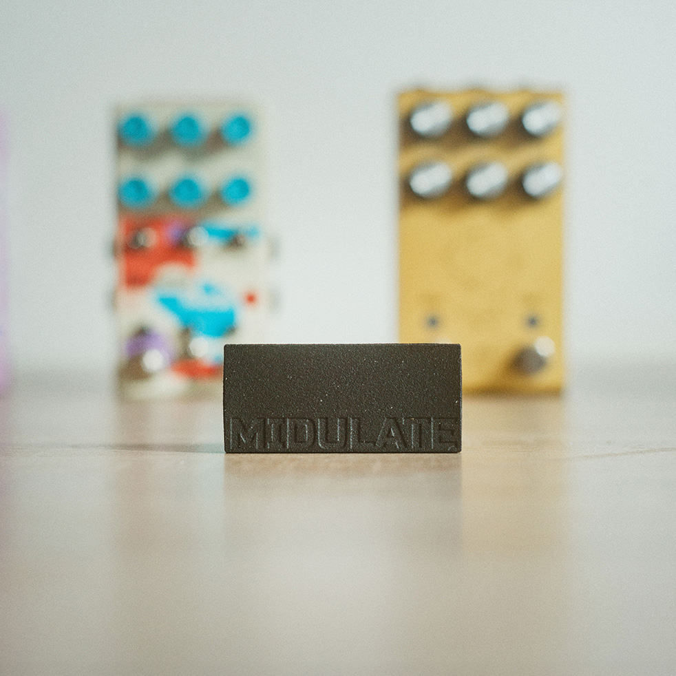 A small black block labeled MODULATE sits on a wooden surface, with two colorful, blurry guitar effect pedals in the background.