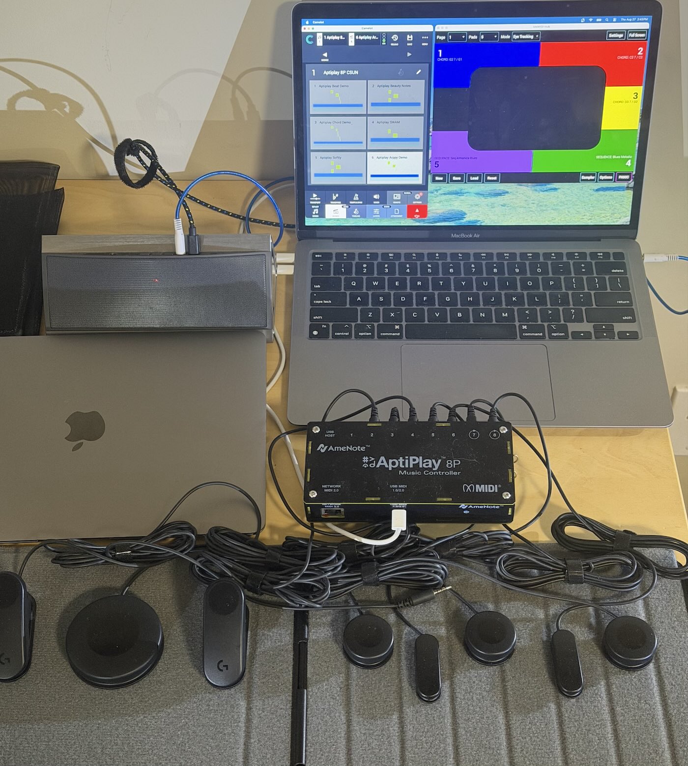 A MacBook with a colorful software interface, a gray Apple laptop, an AptiPlay BP controller, cables, a gray soundbar, and several black input devices arranged on a wooden desk.