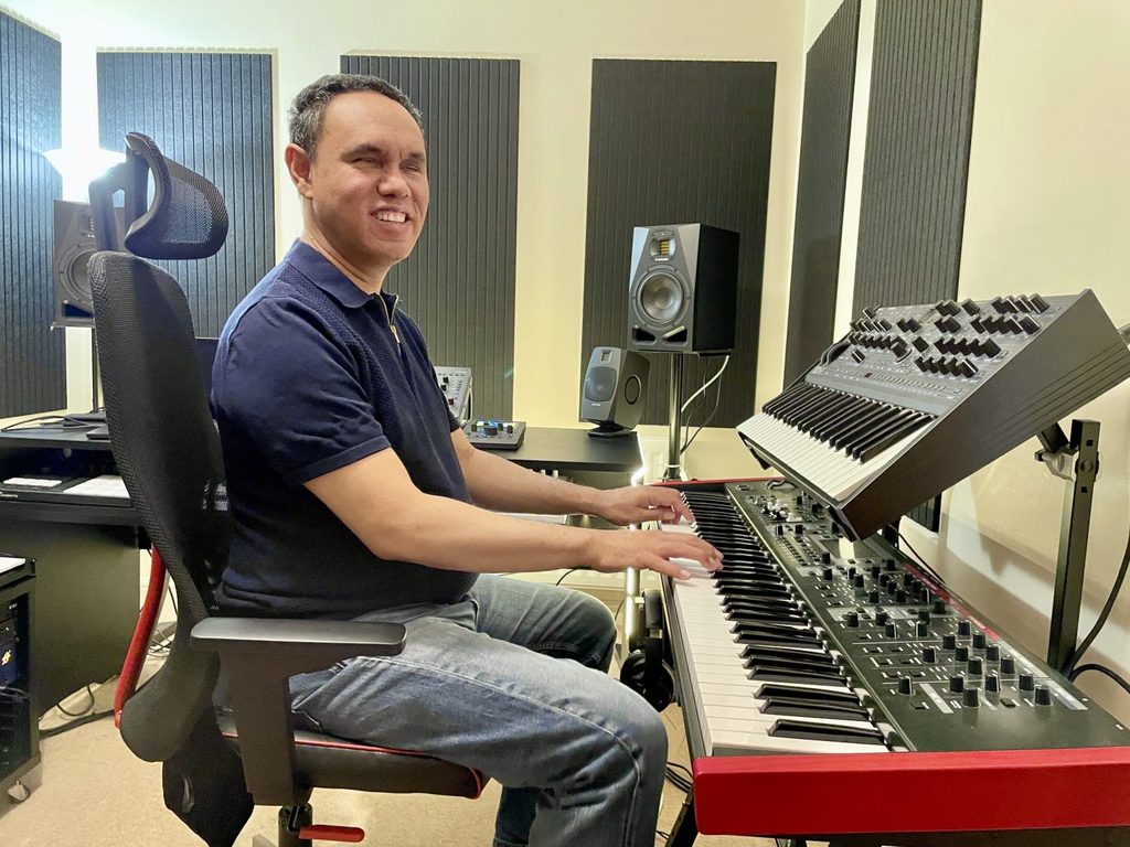 A man smiles while playing a keyboard synthesizer in a music studio, surrounded by audio equipment and soundproof panels.