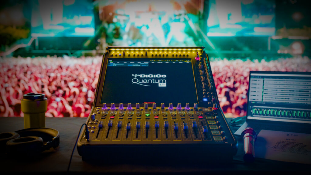 A digital audio mixing console with a DiGiCo Quantum display sits on a desk, with headphones, a coffee cup, a microphone, and a laptop. A large, colorful concert crowd is blurred in the background.