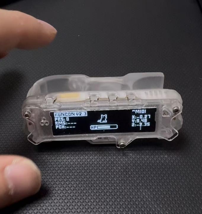 A hand holds a transparent device with a small screen displaying data, icons, and a figure. The device reads PLUGDRV2.3 and shows various numeric values on a black background.