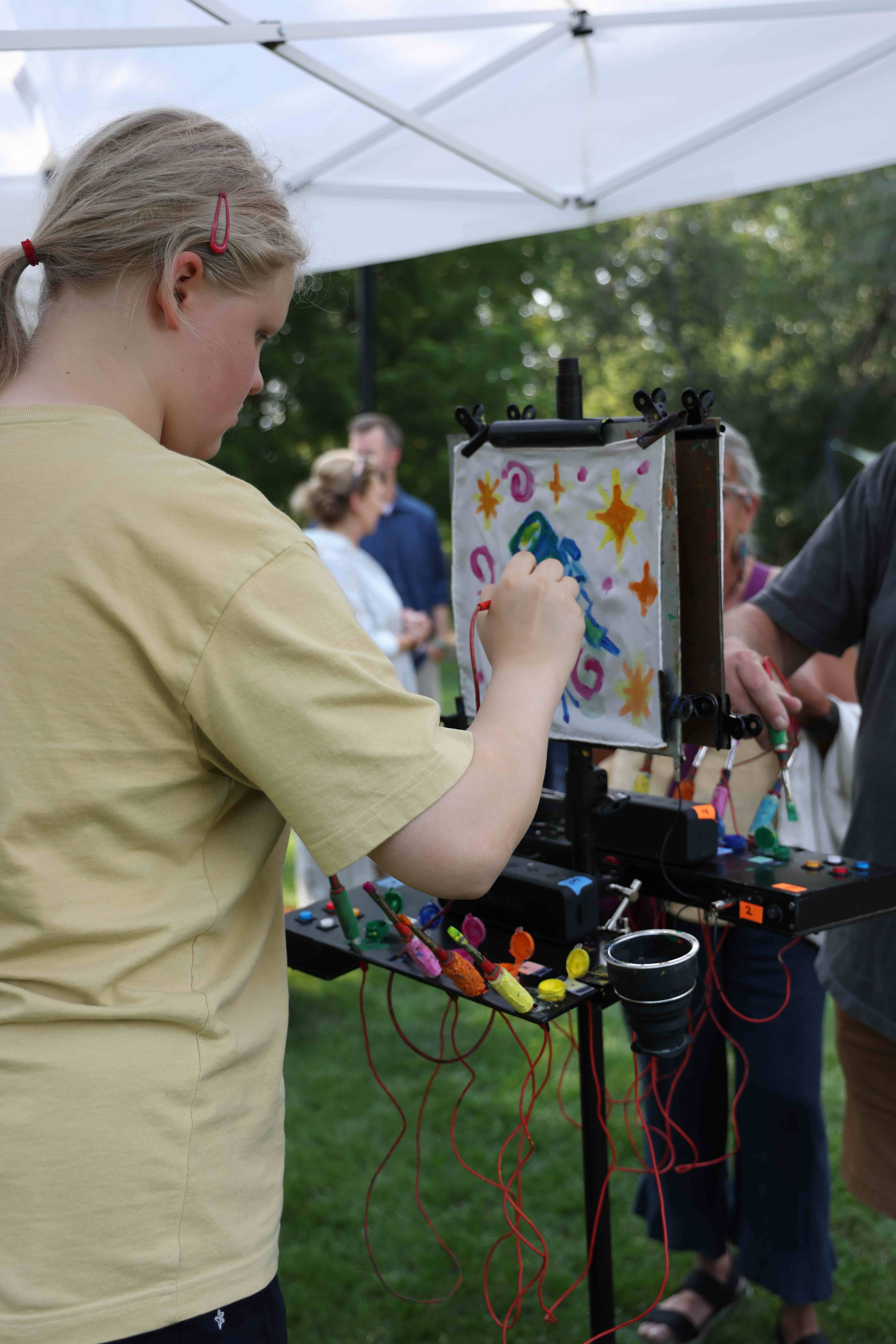 A person in a yellow shirt paints a colorful design on a canvas at an outdoor event, using a setup with lights and wires; people and trees are visible in the background.