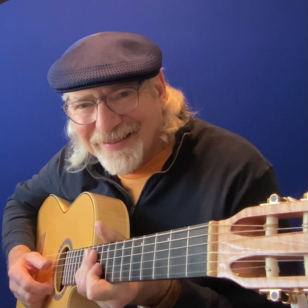 An older man with long gray hair, wearing glasses and a navy flat cap, smiles while playing an acoustic guitar in front of a solid blue background.