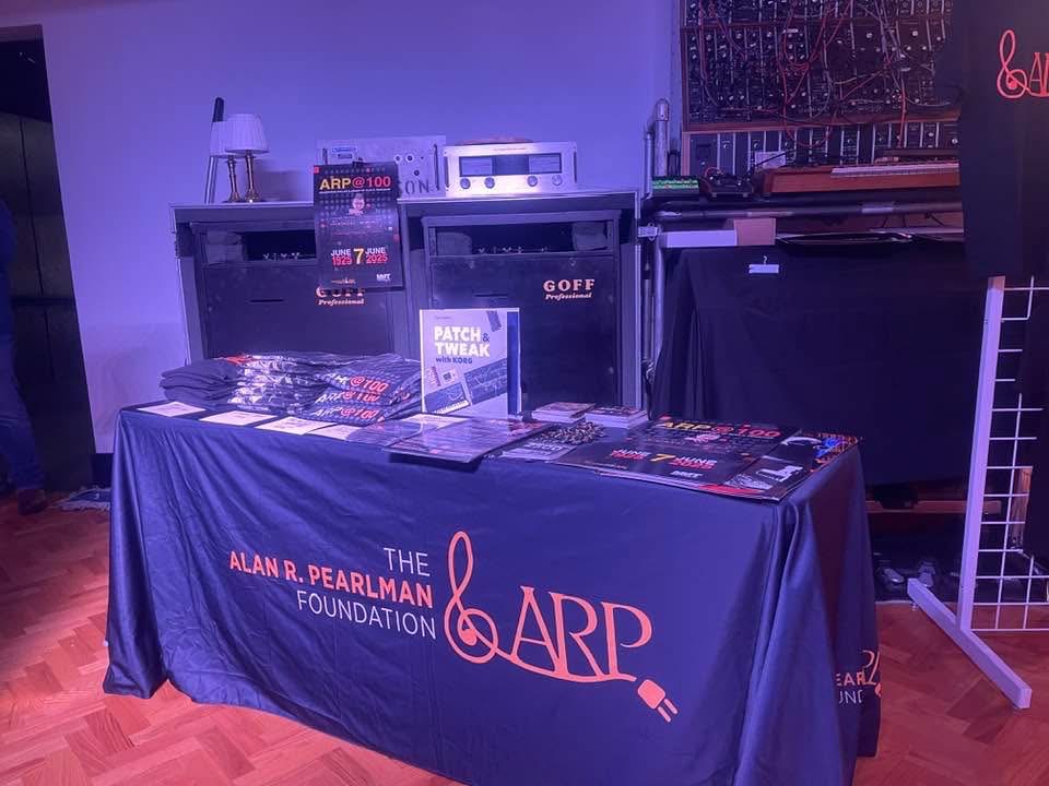 A table covered with The Alan R. Pearlman Foundation cloth displays books, shirts, and magazines. Behind it are electronic equipment racks, a lamp, and a synthesizer in a purple-lit room.