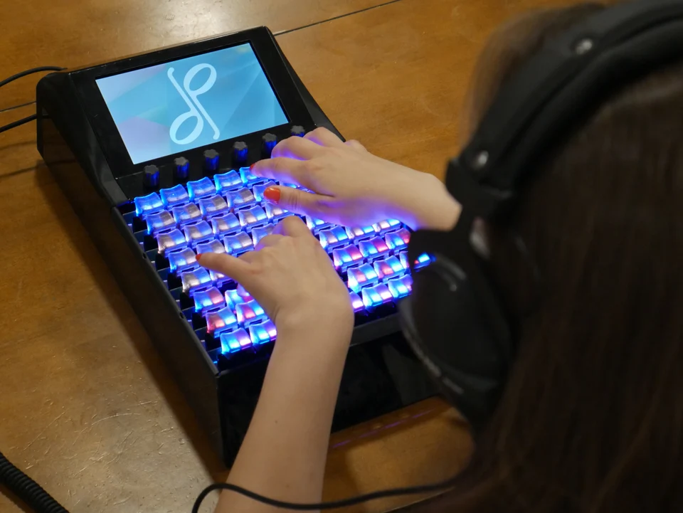 A person wearing headphones plays a light-up electronic music pad with a screen displaying a blue and white logo on a wooden table.