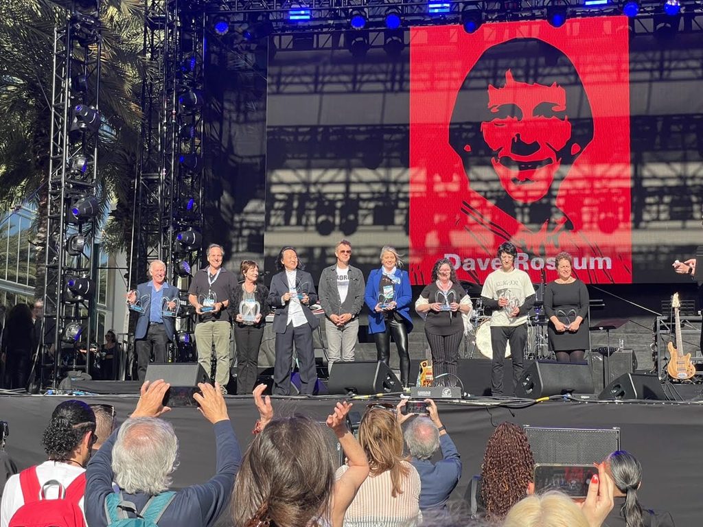 A group of people stand on an outdoor stage holding awards, facing an audience. A large red screen behind them displays an illustrated face and the name Dave Rossum. Palm trees and lighting equipment are visible.