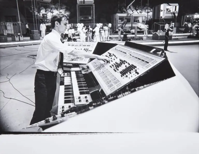 A man in formal attire operates a large, complex control panel with many buttons and switches in a spacious, industrial-looking setting.